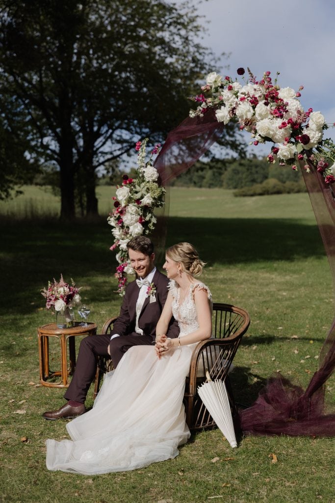 Braut und Bräutigam sitzen unter Blumenbogen bei Hochzeit im Freien.